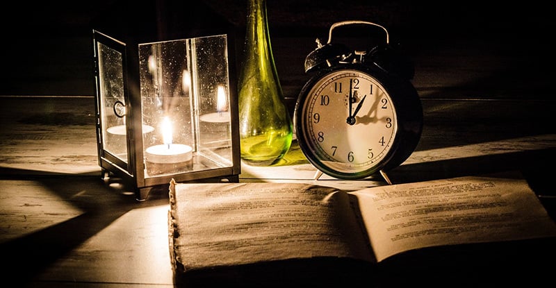 A dimly lit photo featuring a lamp, a clock, and an open book, with a green glass bottle placed between the lamp and the clock.