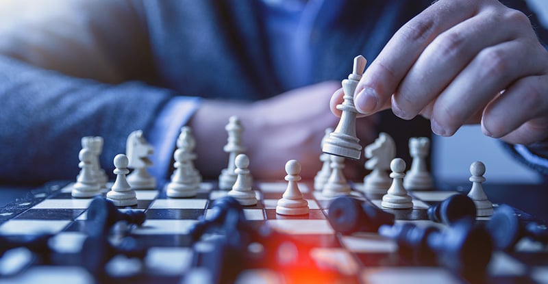 A photo of a chessboard showing a person giving a checkmate to their opponent using a white chess piece. Most of the black pieces are knocked over, while some white pieces remain standing upright.