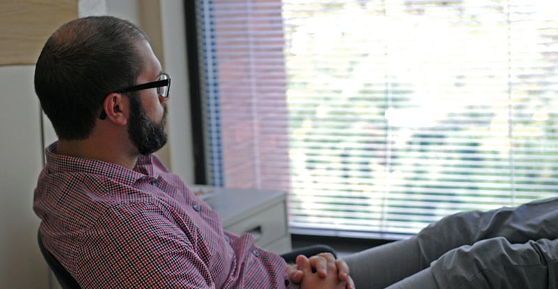 A photo of a man looking outside a window through blinds with his legs resting on his desk and hands folded in his lap.