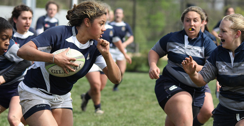 A photo of women playing rugby where one woman is holding the ball while running.