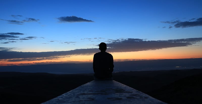 A photo of a person sitting on a ledge watching the sunset.