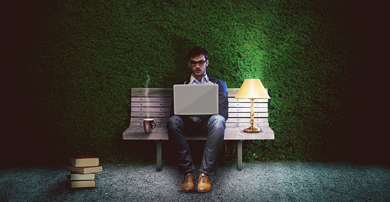 A photo of a person sitting on a bench in front of a tall manicured bush with books on the ground. He can be seen typing on his laptop, a mug on his right and a lamp on his left.