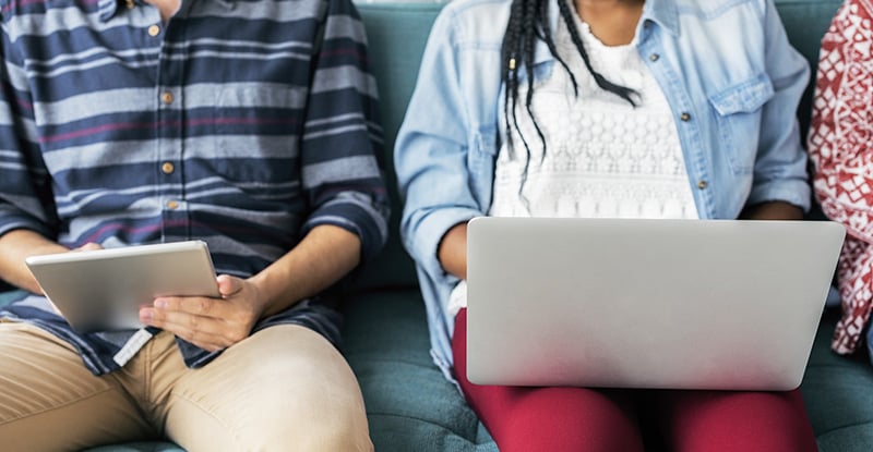 A photo of people seated on a couch, with one person typing on a laptop and another writing on a notepad.