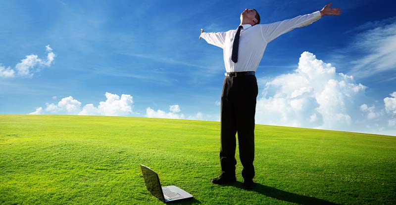 A photo of a man standing with his hands wide open and looking at the sky. There is a laptop on the grass beside his feet to signify freedom.