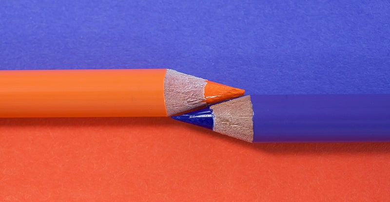 A photo of an orange and a blue colored pencil placed with the sharpened edges against each other, the orange pencil at the top and the blue pencil at the bottom. The background is split with blue at the top and orange at the bottom.