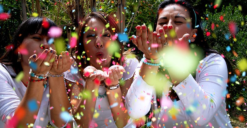 A photo of three women in nature wearing white outfits and blowing confetti toward the camera.