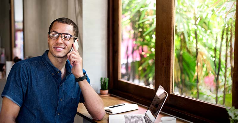 A photo of an ethnic man smiling while on the photo looking away. There is a laptop on the table with a window overlooking a garden.