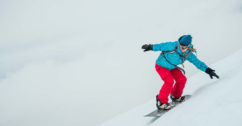 A photo of a woman skiing at the side of a mountain wearing a blue jacket and red ski pants.