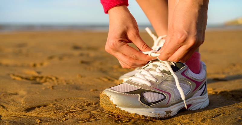 A photo of a person tying their shoe laces on the beach with the sea and sky in the background.