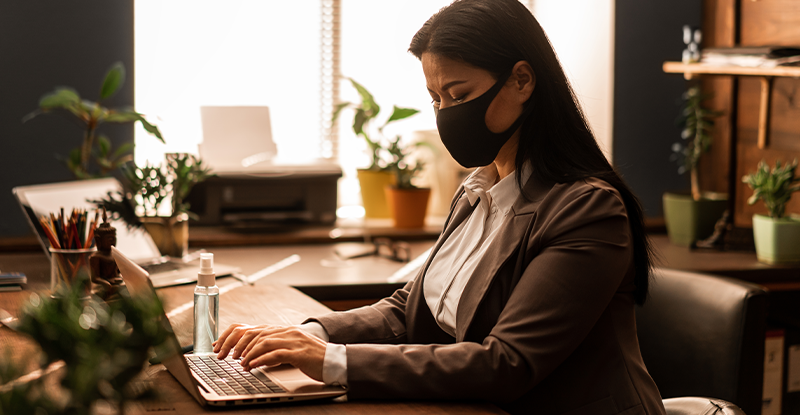 A photo of a professionally dressed Asian woman wearing a face mask while working on her laptop in an office setting surrounded by potted plants and stationery.