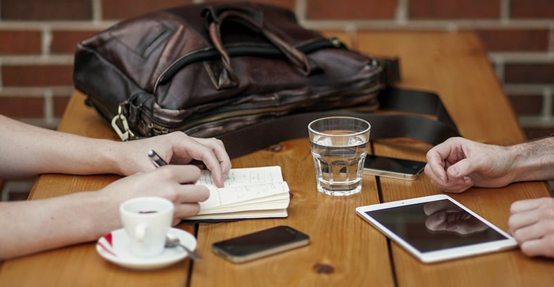 A photo of a desk with two people sitting on either side; one person is writing in a notebook, and the other has their hands resting on the table.