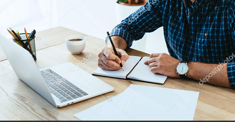 A Caucasian man wearing a blue and black checked shirt, writing on a notebook while looking at his laptop.