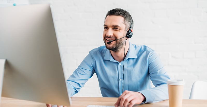 A Caucasian man with a headset smiling and looking at his computer. There is a coffee cup on the right side of the desk.