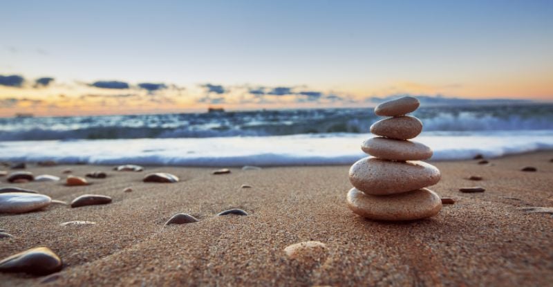 A photo of flat circular stones stacked on top of one another on the sand against a backdrop of the sea and blue sky.