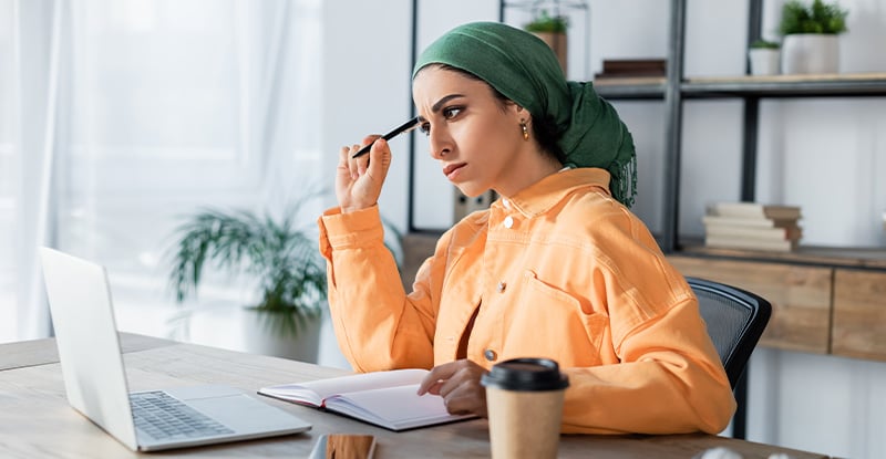 A photo of an Arab woman wearing a green turban and an orange shirt, thinking with a pencil to her head while looking at her laptop. She is seated in an office with a shelf in the background holding books and potted plants.