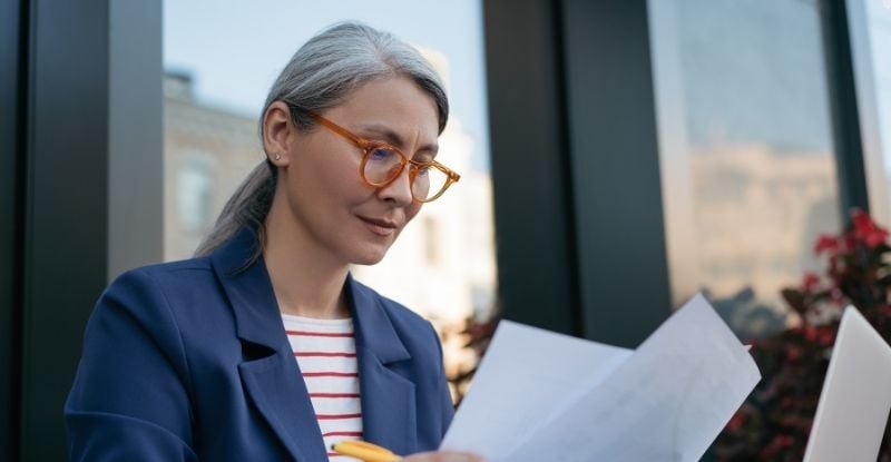 An Asian woman with yellow-framed glasses holds a stack of papers in one hand and a pen in the other. She is looking at the papers intently, with the backdrop of an office building window.