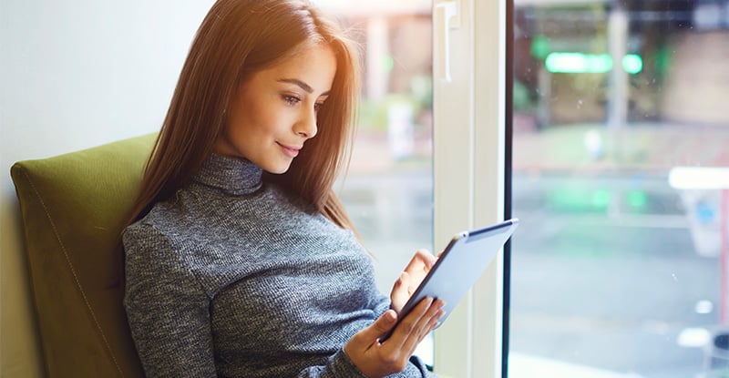 A Caucasian woman is lounging on a couch and looking at a tablet in her hands.