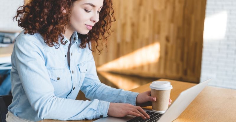 A photo of a woman in an office setting typing on her laptop while holding a coffee cup in her left hand.