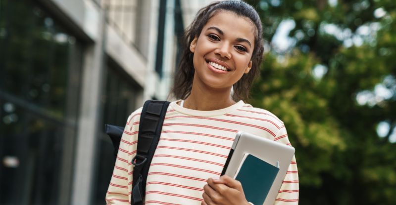 A photo of an African student wearing an orange horizontally striped shirt and carrying a backpack, smiling at the camera. In one hand, they hold a laptop and a notebook. The background features a university building and trees, creating an academic atmosphere.