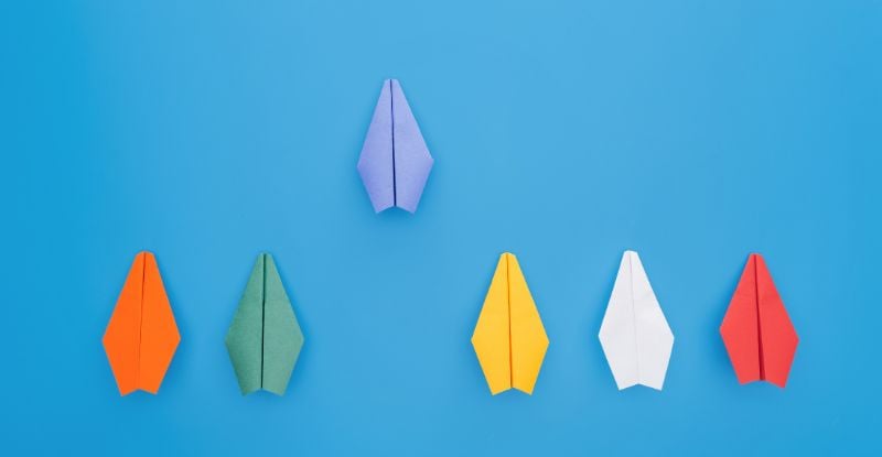 An illustration against a blue background featuring six paper airplanes, with one airplane positioned ahead to represent progress and getting ahead in life.