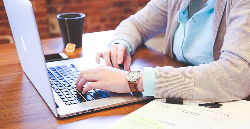 A photo of a woman typing on a laptop with a notebook on one side and a tea mug on the other.