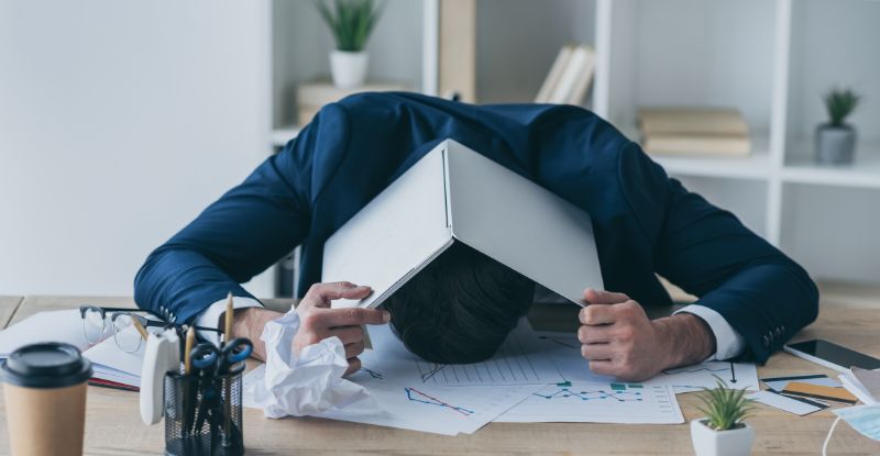 A photo of a man with his head on the table and a laptop over his over with a paper crumpled near by signifying frustration. 