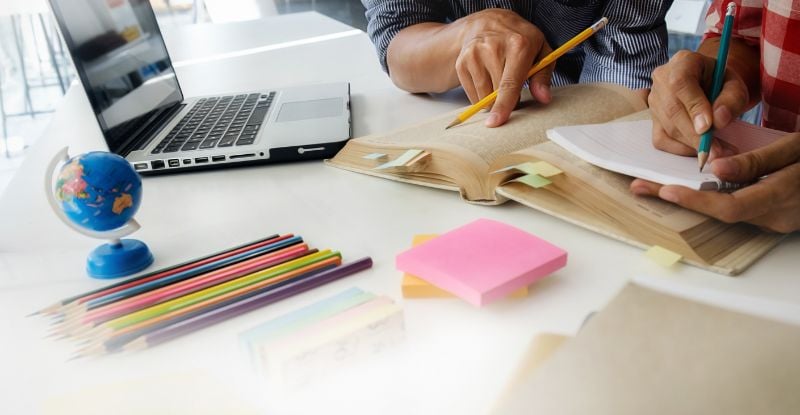 A photo of two people, where one is reading from a book and the other is taking notes. There is a laptop, globe, sticky notes, colour pencils and papers placed around their book on the table.