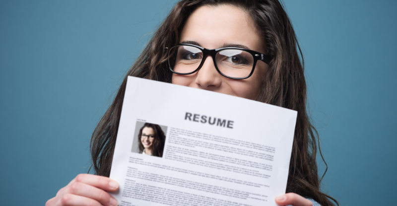 A photo of a woman wearing glasses holding up her resume with her picture on it. The photo is taken against a blue background.