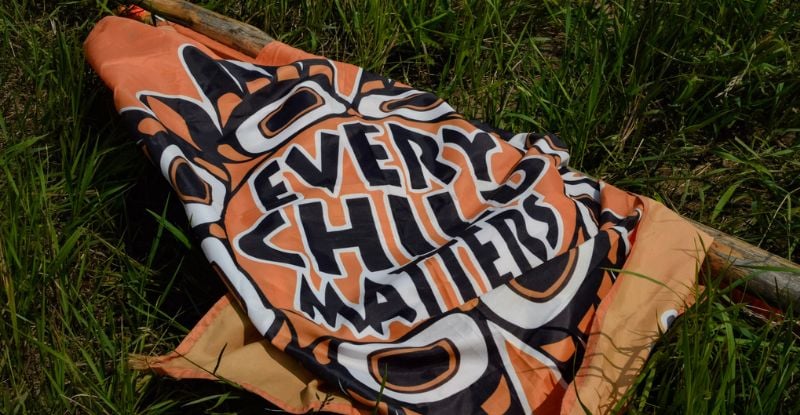 A photo of an orange cloth on the grass that reads "Every child matters" with Indigenous symbols around the text in black and white.