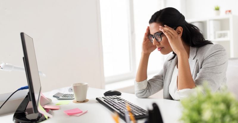 A woman wearing a suit and glasses looks frustrated with her on either side of her head, while looking at her computer. There are sticky notes, coffee mug and a calculator on the table.