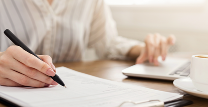 A photo of a woman writing on a notebook while having a finger on the trackpad of her laptop.