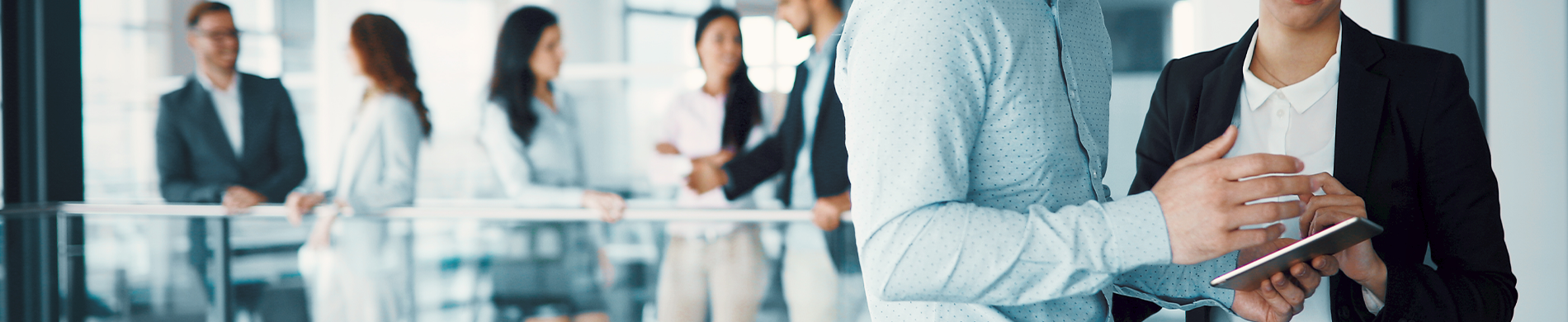 Banner A man and a woman talking, with the man holding a tablet, while other people are conversing in the background in an office setting.