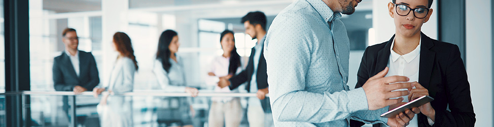 Mobile Banner A man and a woman talking, with the man holding a tablet, while other people are conversing in the background in an office setting.